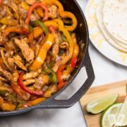 A cast iron skillet filled with chicken fajitas with tortillas and garnishes alongside.