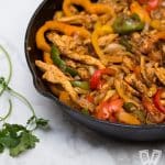 A cast iron skillet filled with chicken fajitas with tortillas and garnishes alongside.