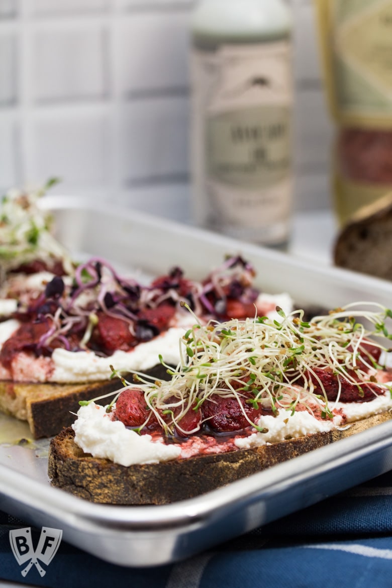 ¾ view of 3 pieces of toast topped with 3 types of sprouts, balsamic-roasted strawberries and ricotta with ingredients in the background.