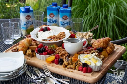 3/4 view of a board of assorted breakfast items sitting on an outdoor table with dishes and beverages around.