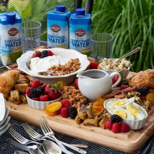 ¾ view of a board of assorted breakfast items sitting on an outdoor table with dishes and beverages around.