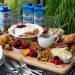 3/4 view of a board of assorted breakfast items sitting on an outdoor table with dishes and beverages around.