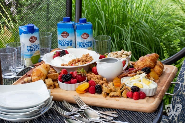 ¾ view of a board of assorted breakfast items sitting on an outdoor table with dishes and beverages around.