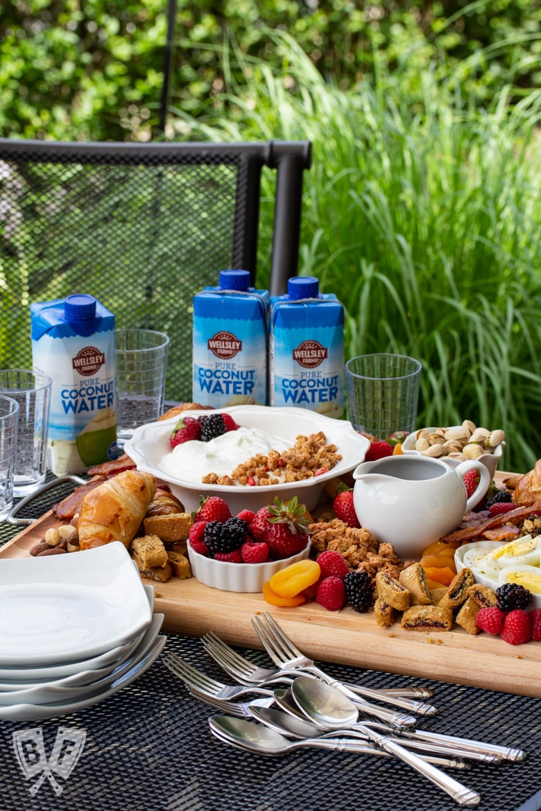 ¾ view of a board of assorted breakfast items sitting on an outdoor table with dishes and beverages around.