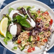 Overhead view of a plate of Mediterranean Farro Salad with Spiced Beef Patties drizzled with yogurt over greens served with lemon wedges and fresh herbs.