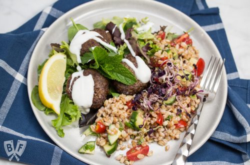 Overhead view of a plate of Mediterranean Farro Salad with Spiced Beef Patties drizzled with yogurt over greens served with lemon wedges and fresh herbs.