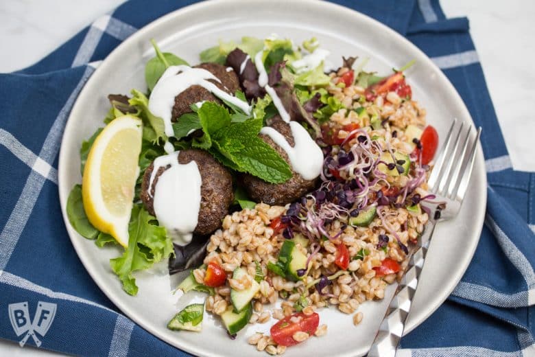 Overhead view of a plate of Mediterranean Farro Salad with Spiced Beef Patties drizzled with yogurt over greens served with lemon wedges and fresh herbs.