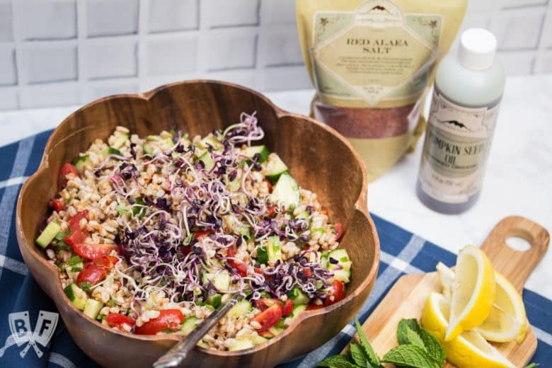 Overhead view of Mediterranean Farro Salad, with ingredients in the background and a cutting board of lemon wedges with fresh herbs.