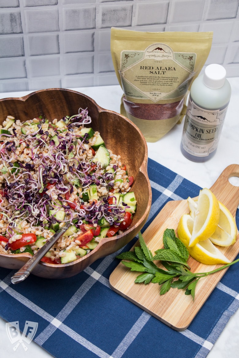 Overhead view of Mediterranean Farro Salad, with ingredients in the background and a cutting board of lemon wedges with fresh herbs.