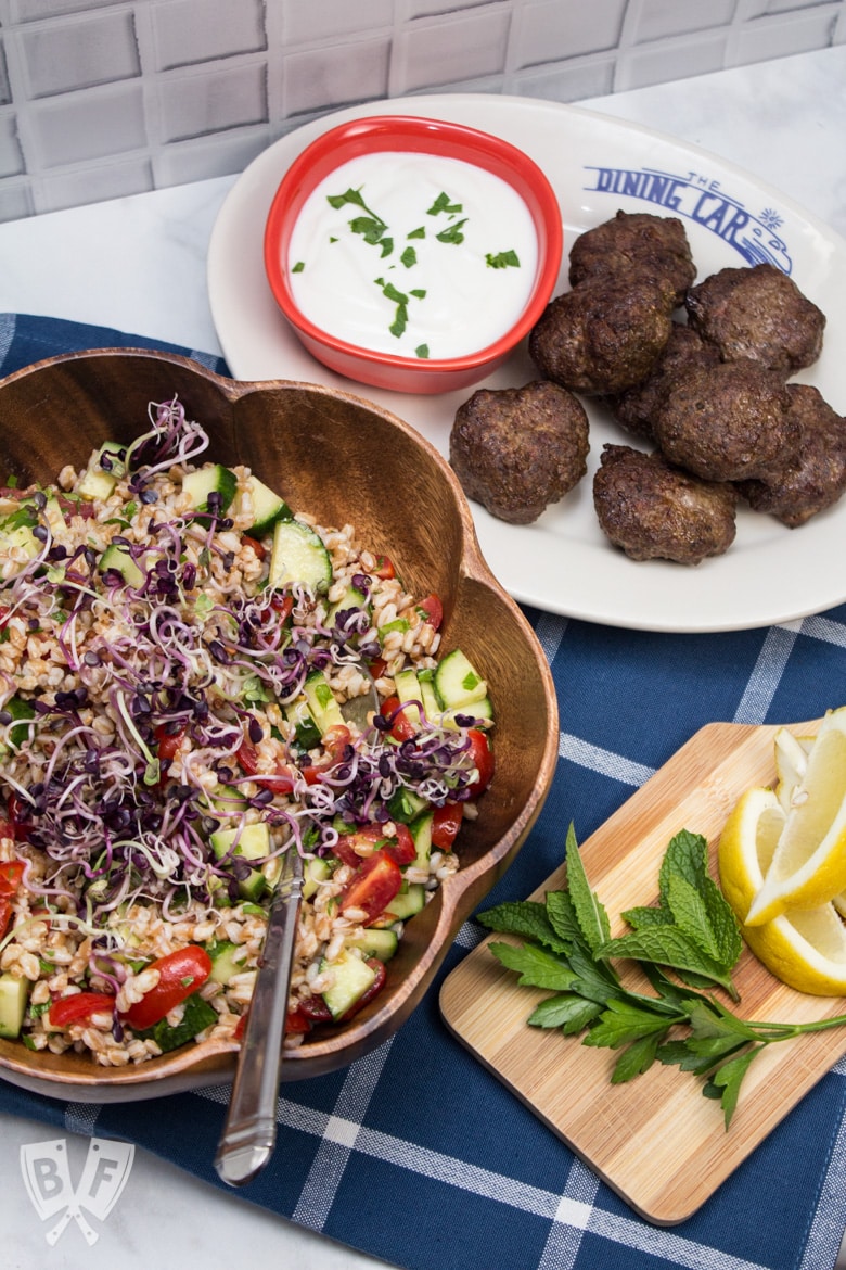 Overhead view of Mediterranean Farro Salad with Spiced Beef Patties and a cutting board of lemon wedges with fresh herbs.