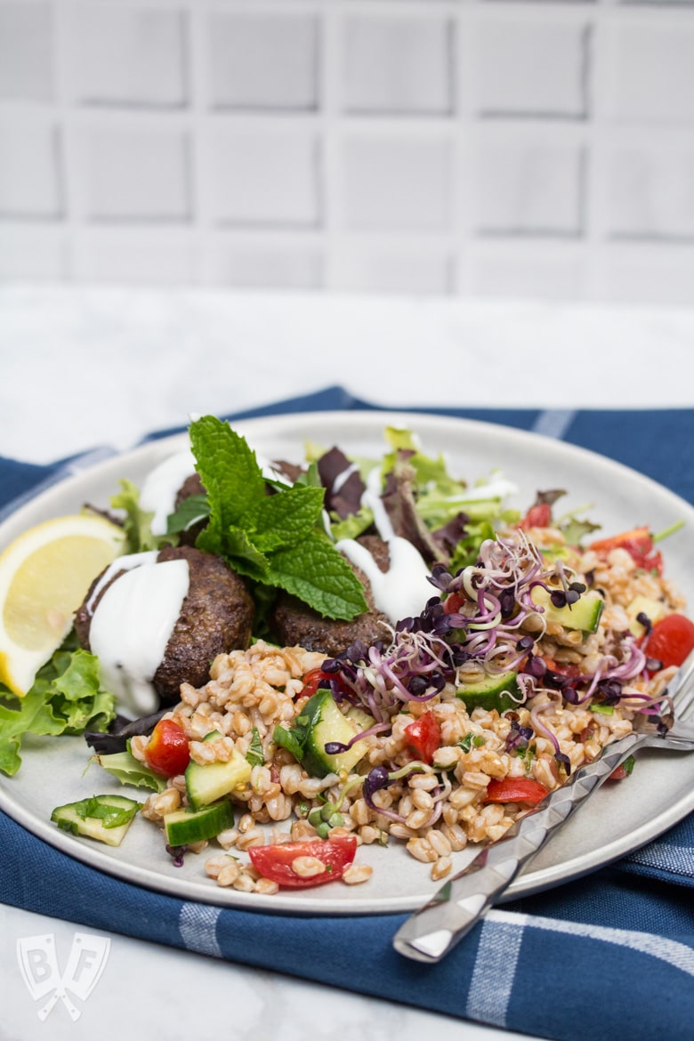 ¾ view of a plate of Mediterranean Farro Salad with Spiced Beef Patties drizzled with yogurt over greens served with lemon wedges and fresh herbs.