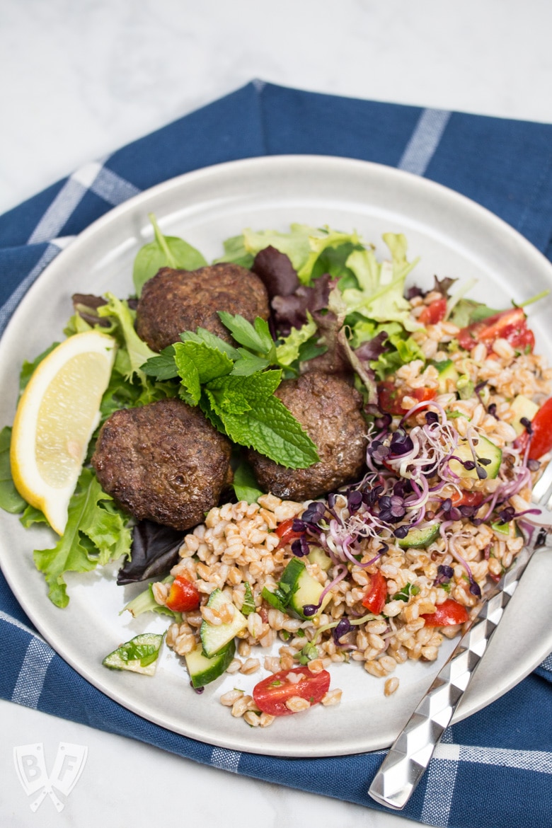 Overhead view of a plate of Mediterranean Farro Salad with Spiced Beef Patties over greens served with lemon wedges and fresh herbs.