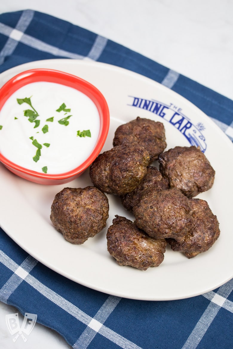 Overhead view of a plate of spiced beef patties with a bowl of yogurt and fresh herbs