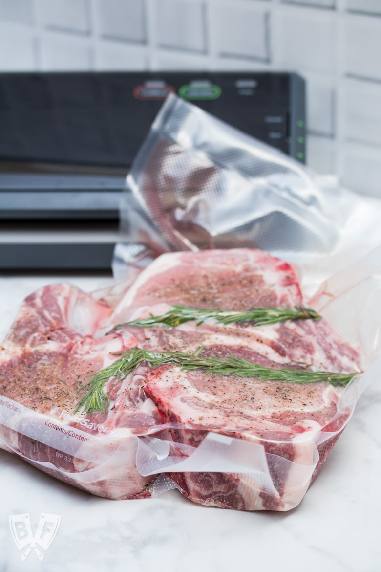 A vacuum-sealed bag of pork chops and rosemary with a FoodSaver machine in the background.