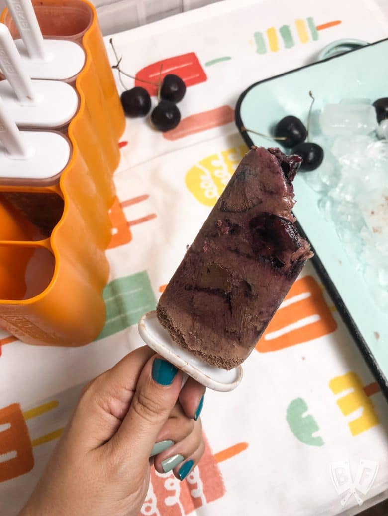 A hand holding a Black Forest Popsicle next to a tray of ice and a popsicle maker.