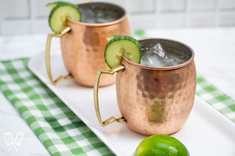 ¾ view of 2 copper mugs filled with a Cucumber Moscow Mule cocktails with ingredients alongside.