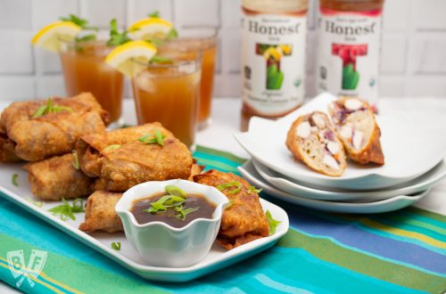3/4 view of a platter of General Tsao’s Chicken Egg Rolls with a bowl of dipping sauce and glasses and bottles of iced tea.