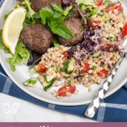 Overhead view of a plate of Mediterranean Farro Salad with Spiced Beef Patties over greens served with lemon wedges and fresh herbs.