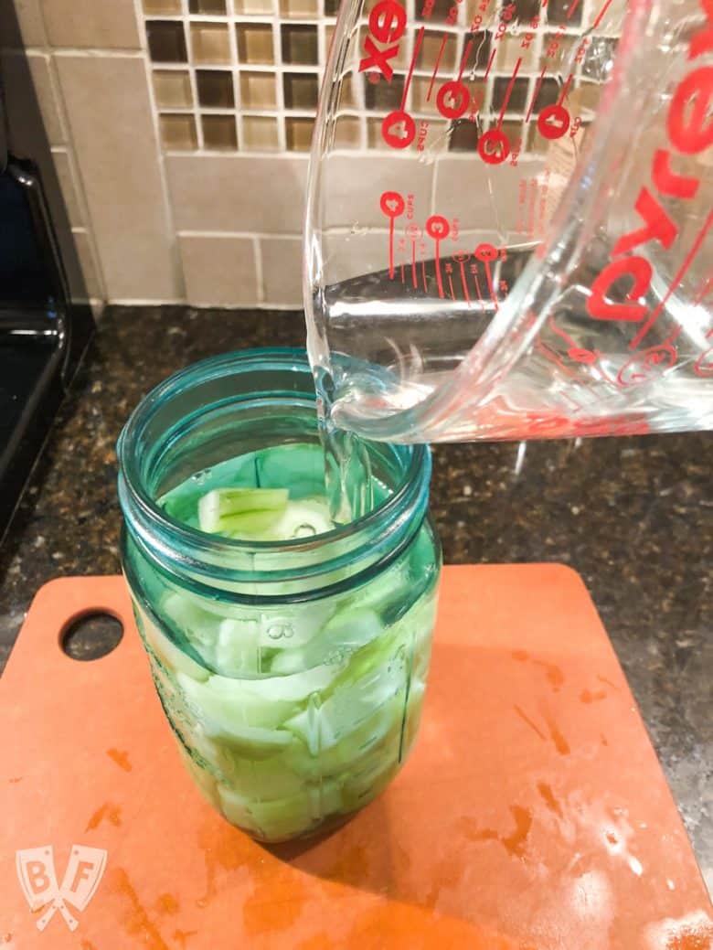 ¾ view of pouring vodka into a jar of cucumbers for infusing.