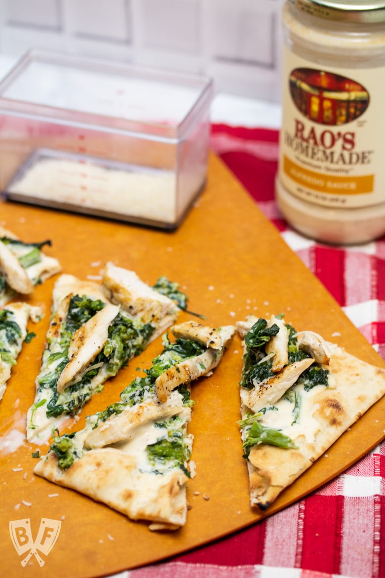 Chicken and broccoli rabe pizza cut into slices with Parmesan cheese and a jar of alfredo sauce in the background.