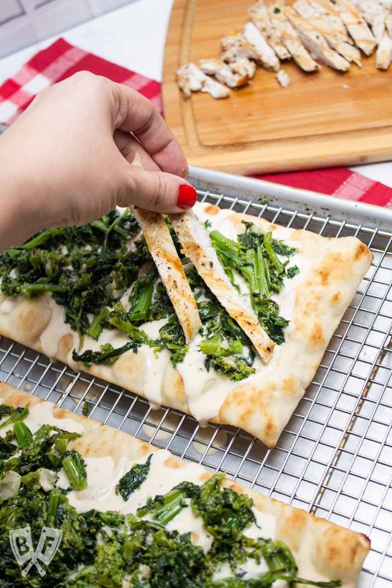 Hand placing grilled chicken onto pizza crust with alfredo sauce and broccoli rabe.