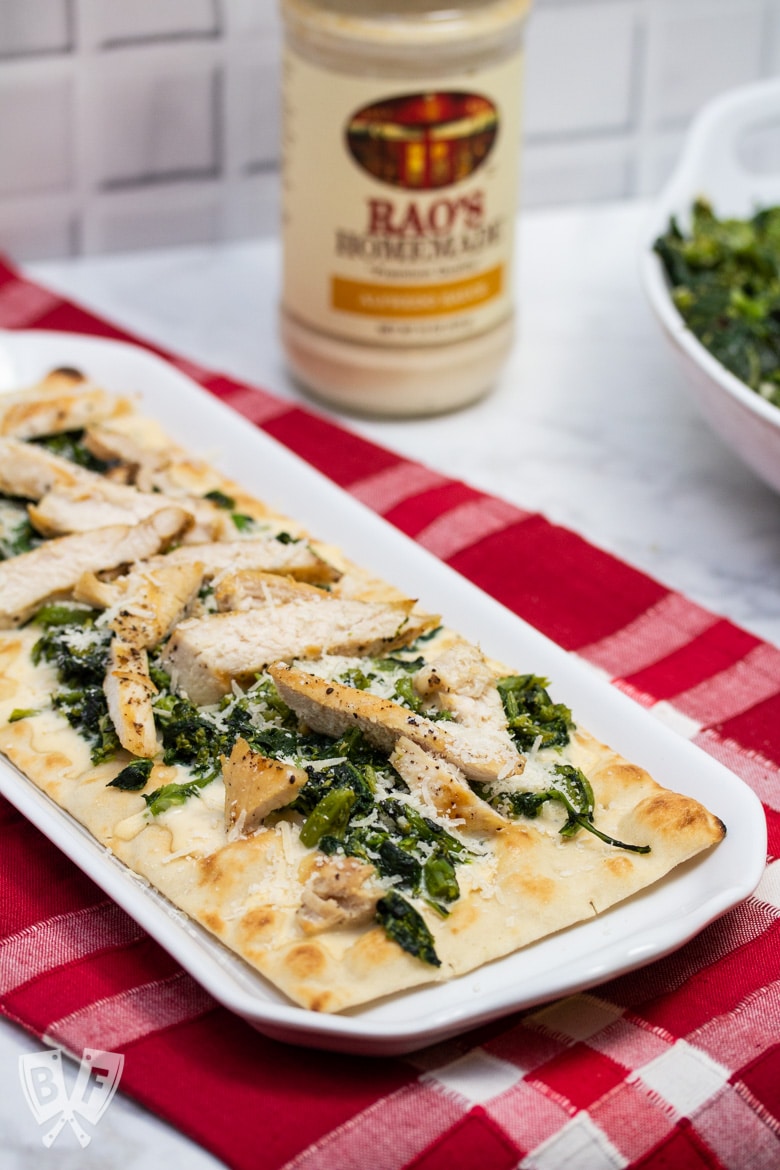 Platter of chicken and broccoli rabe pizza with a jar of alfredo sauce and bowl of broccoli rabe in the background.