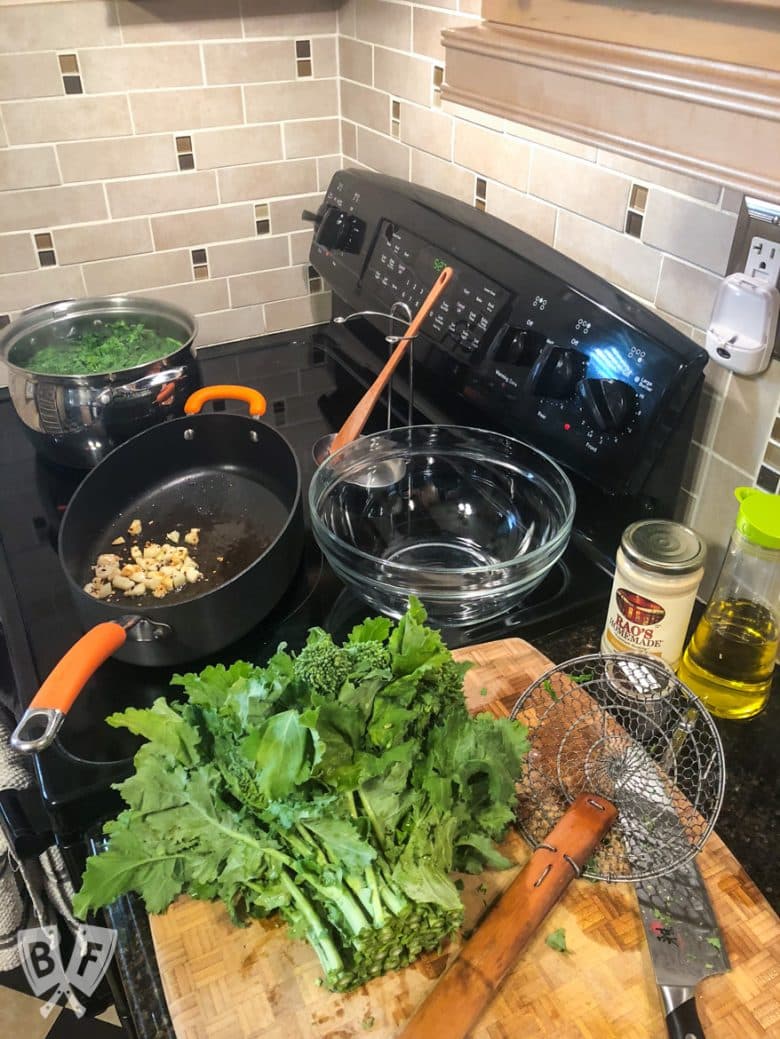 Broccoli rabe on a cutting board with a skillet of garlic and a pot of boiling water in the background.