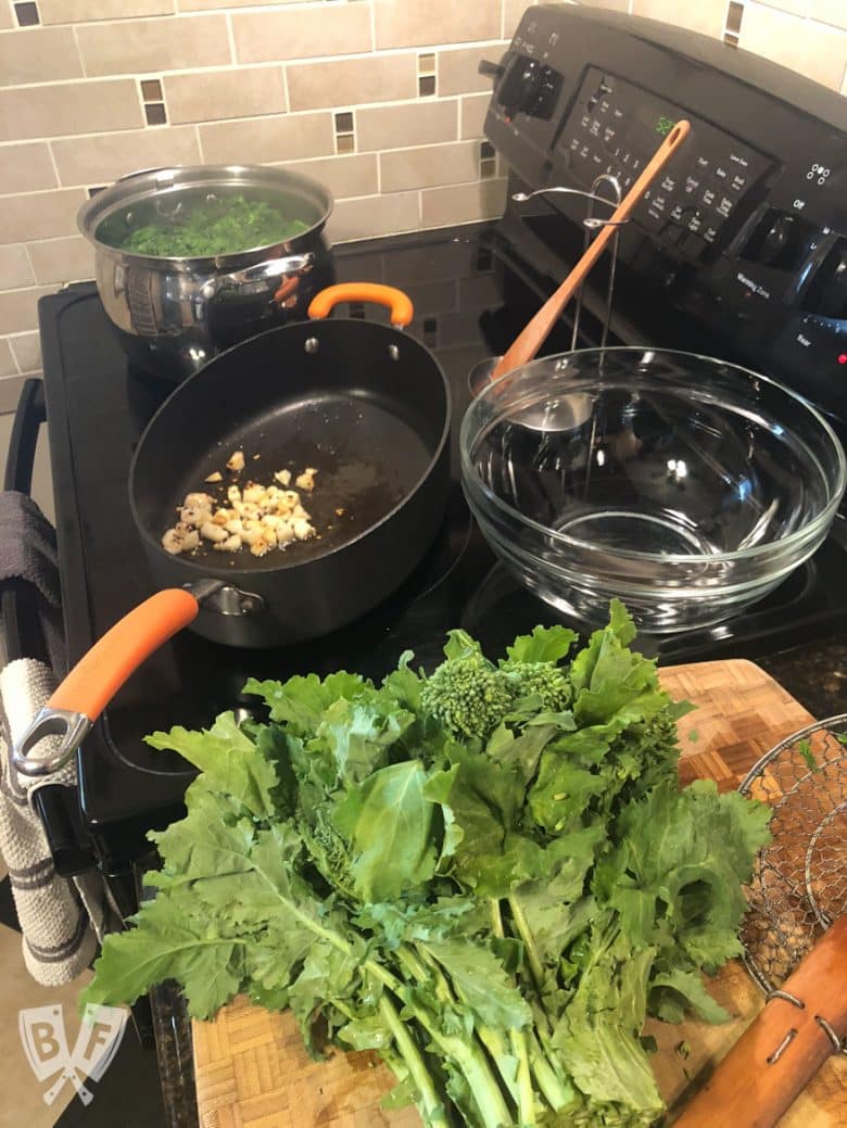 Broccoli rabe on a cutting board with a skillet of garlic and a pot of boiling water in the background.