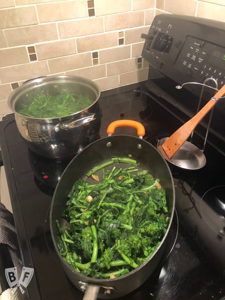 Broccoli rabe on a cutting board with a skillet of garlic and a pot of boiling water in the background.