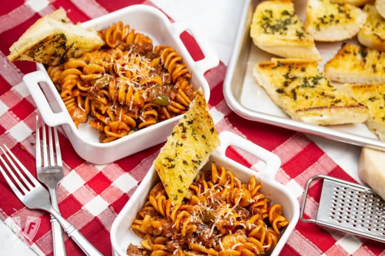 Overhead view of dishes of Instant Pot Tomato Beef Pasta with serving dishes with Parmesan cheese and a tray of garlic bread in the background.