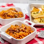 3/4 view of dishes of Instant Pot Tomato Beef Pasta with serving dishes with Parmesan cheese and a tray of garlic bread in the background.