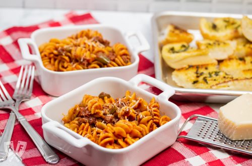 3/4 view of dishes of Instant Pot Tomato Beef Pasta with serving dishes with Parmesan cheese and a tray of garlic bread in the background.