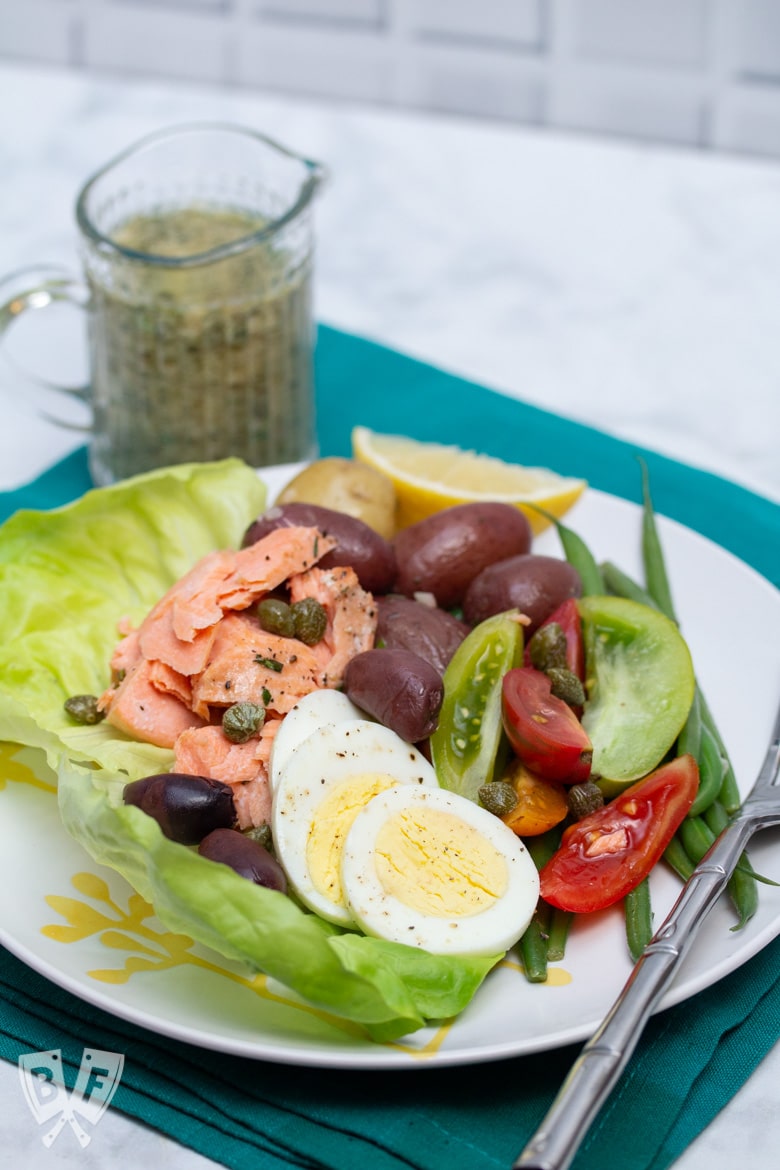 3/4 view of a plate of King Salmon Niçoise Salad with a pitcher of tarragon vinaigrette alongside.