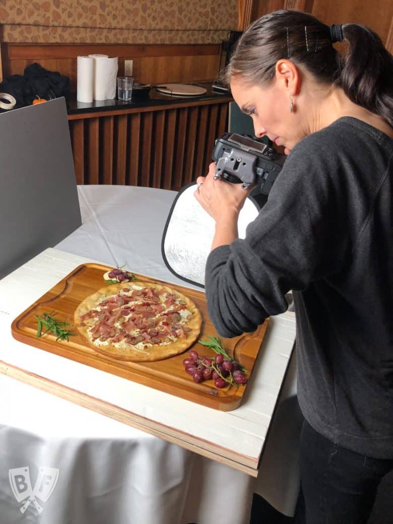 A photographer shooting photos of a burrata pizza with prosciutto and pickled grapes.