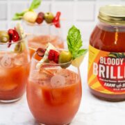 Side view of 3 Bloody Mary cocktails garnished with Italian antipasto ingredients next to a jar of Bloody Mary mix.