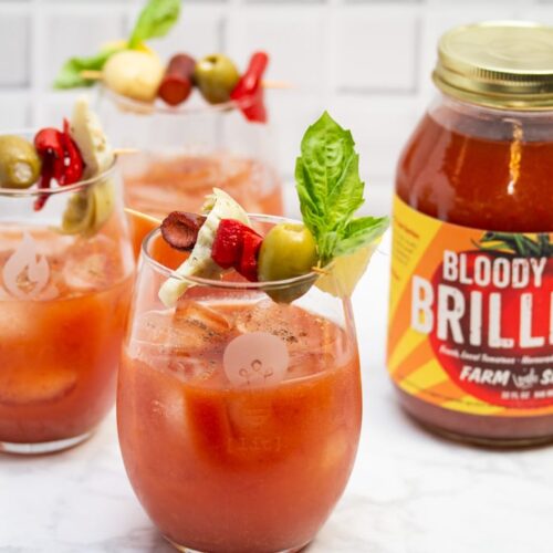 Side view of 3 Bloody Mary cocktails garnished with Italian antipasto ingredients next to a jar of Bloody Mary mix.