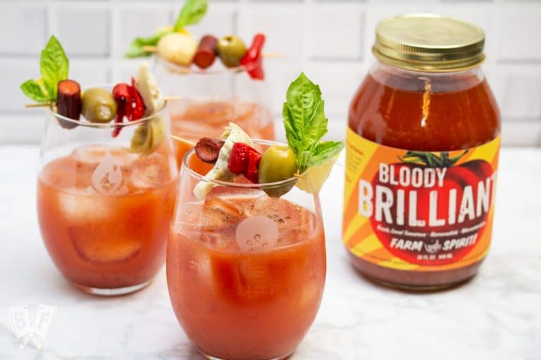 Side view of 3 Bloody Mary cocktails garnished with Italian antipasto ingredients next to a jar of Bloody Mary mix.