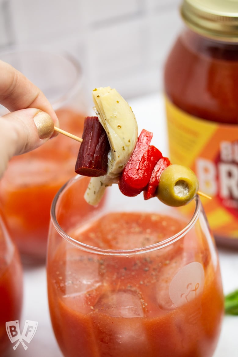 Hand placing a skewer of Italian antipasto ingredients on top of a Bloody Mary cocktail.