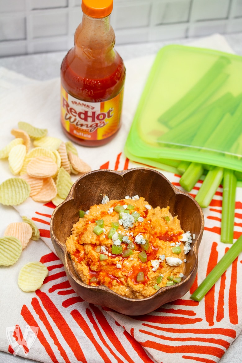 Buffalo chickpea dip with veggie chips, celery sticks, and a bottle of wing sauce.