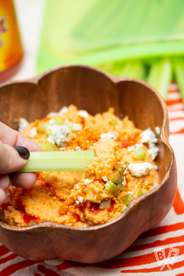 Dipping a celery stick in a bowl of buffalo chickpea dip