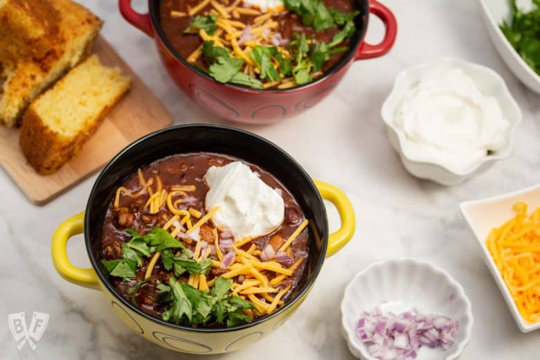 Overhead view of bowls of Spicy Turkey Three-Bean Chili with toppings and cornbread alongside.