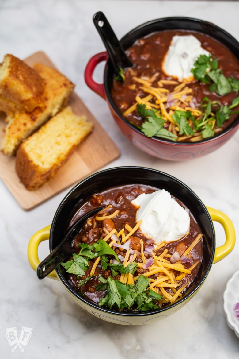 Overhead view of bowls of Spicy Turkey Three-Bean Chili with toppings and cornbread alongside.