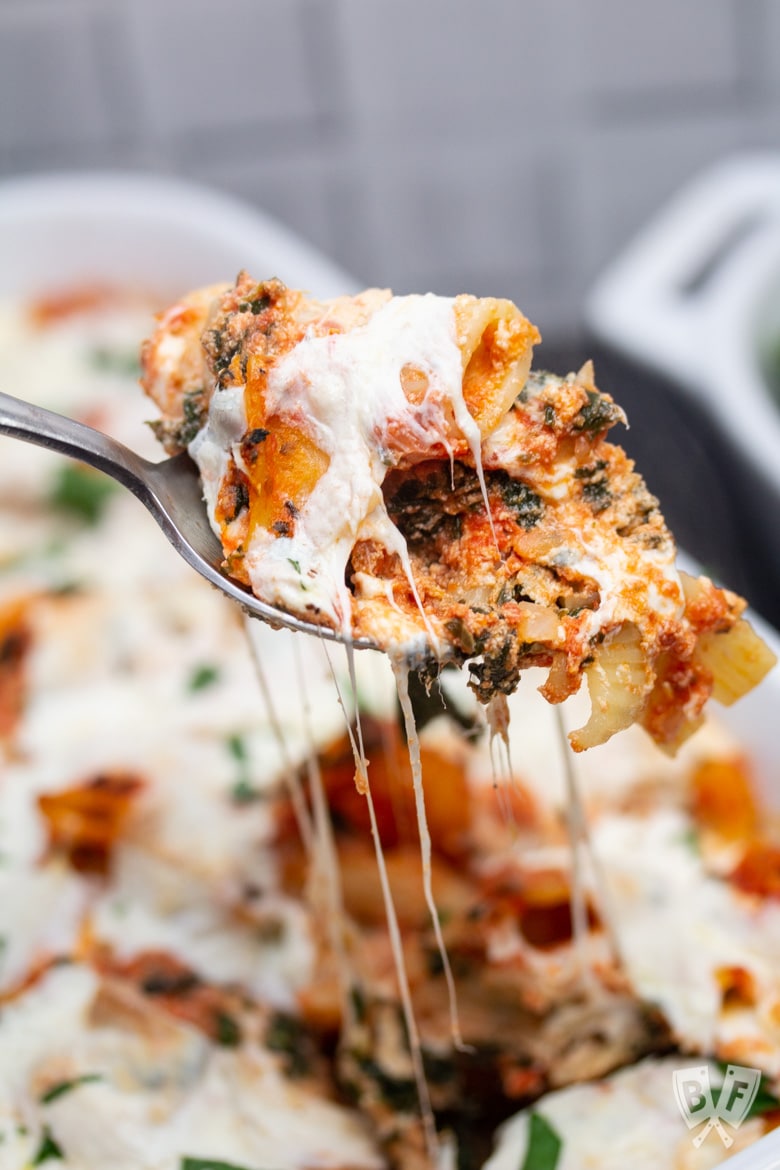 Spoon pulling a serving of baked rigatoni out of a casserole dish.