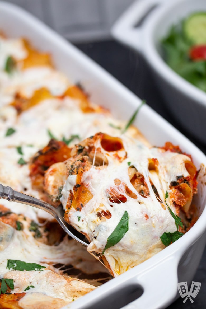 Spoon pulling a serving of baked rigatoni out of a casserole dish.