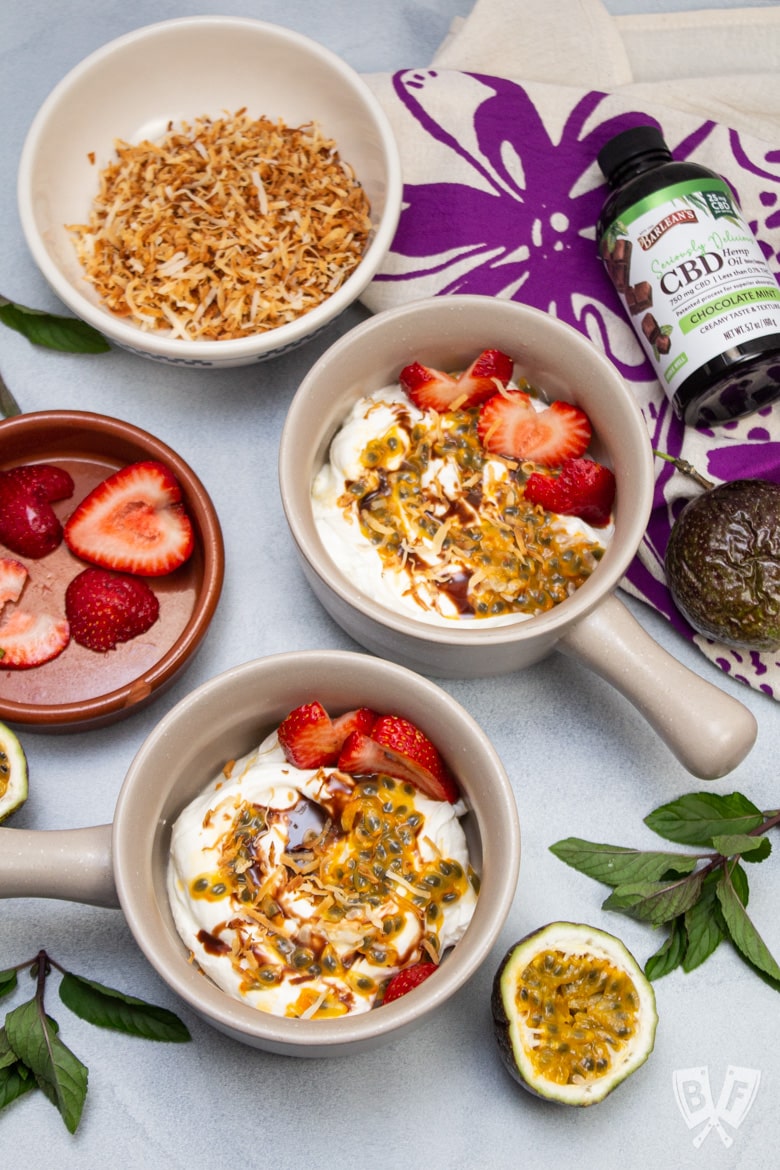 Overhead view of Greek yogurt parfaits with colorful tropical fruit toppings with a bottle of CBD hemp oil alongside.