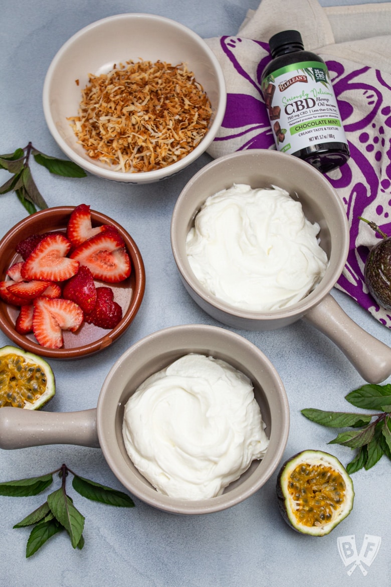 Overhead view of bowls of Greek yogurt with tropical fruit and chocolate mint CBD oil alongside.