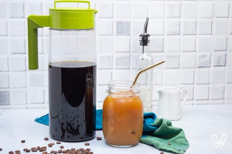 A glass of cold brew coffee with a pitcher, milk, and sweetener alongside.