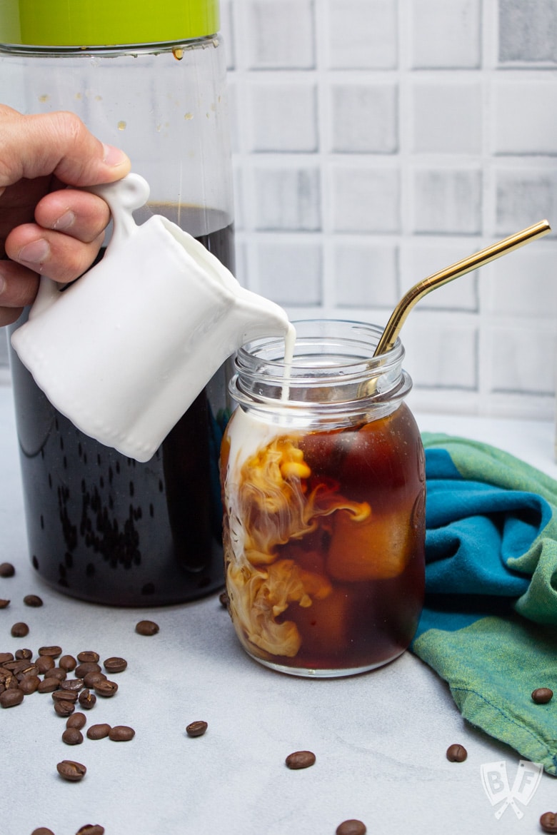 Pouring milk from a pitcher into a glass of homemade cold brew coffee.