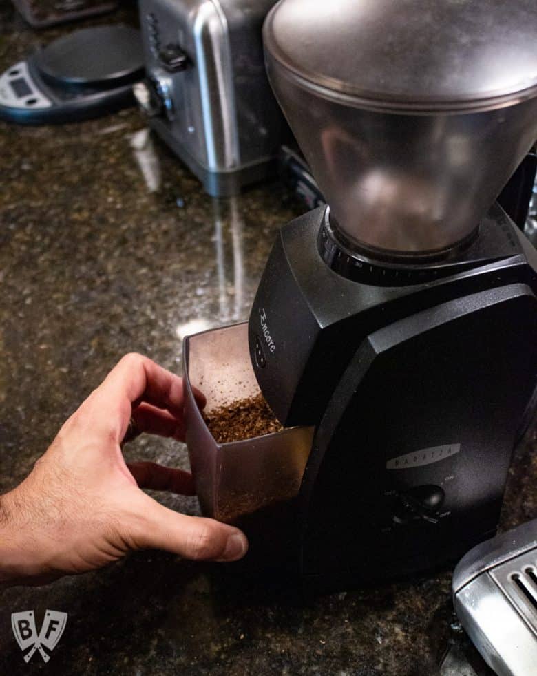 Removing a container of freshly ground coffee beans from a burr grinder to make cold brew coffee