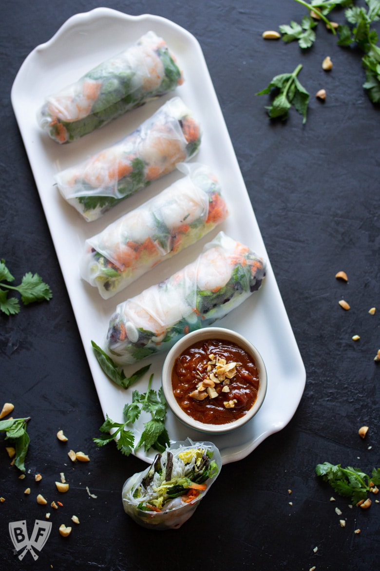 Platter of Vietnamese fresh spring rolls with shrimp and peanut dipping sauce and ingredients sprinkled around.